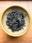 A crackle glazed bowl holding large, wiry Jin Xuan black tea leaves on a wooden background